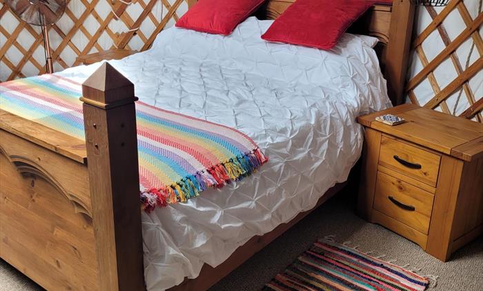 Chunky carved wood bed with white pintuck cotton bedding, a bright rainbow coloured stripy blanket and red velvet cushions, wood bedside cabinet and b